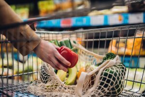 The hand of a person reaching into a grocery cart placing a red bel pepper in their reusable bag.