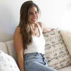 Photo of writer Sam Negrin. She's wearing a white tank top and jeans and is sitting on a beige couch with decorative pillows.