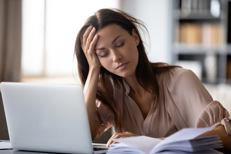 A brunette woman leaning on her hand sleeping in front of an open book and computer.