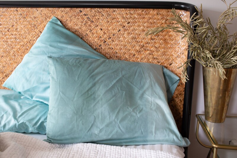 Three ombre tie-dyed pillowcases arranged casually on a bed against a woven headboard. A plant in a vase sits on the nightstand beside the bed. 