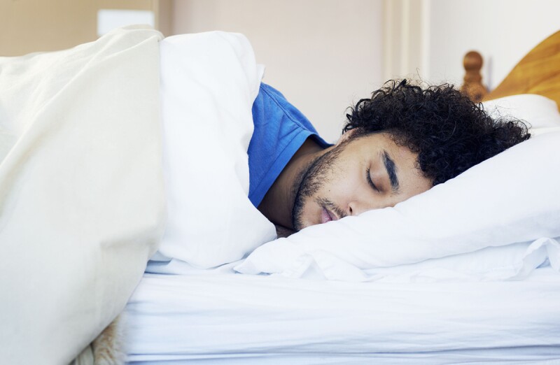 A man in a blue shirt rests his head on a pillow in a bed with white sheets and linens. Right now, sleep matters more than ever. 