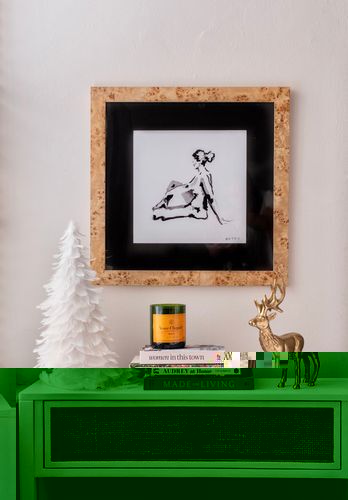 Guest bedroom decor for the holidays. A framed picture of a woman hangs above a nightstand dressed with a petit faux Christmas tress, a stack of books with a candle on top, and a reindeer. 