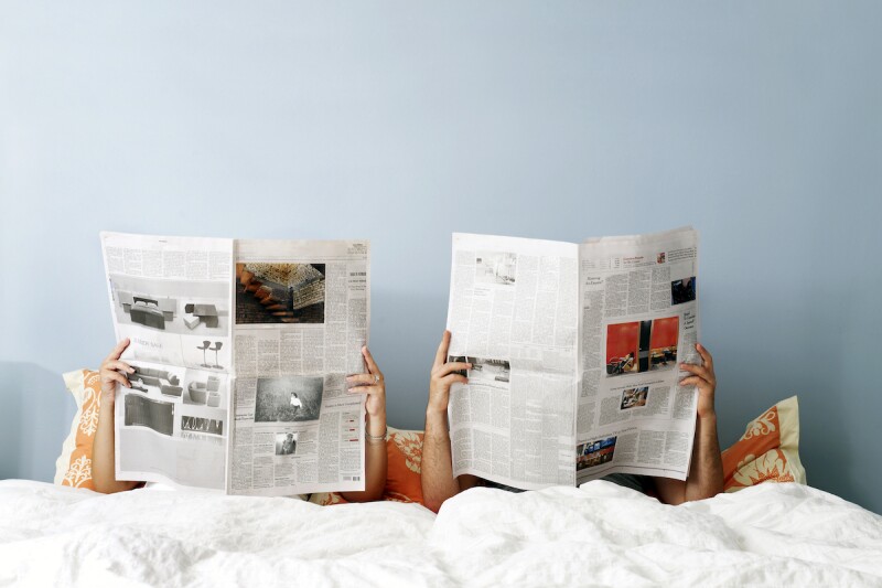 Two people lying in bed in a blue bedroom, each holding open newspapers that block their faces. 