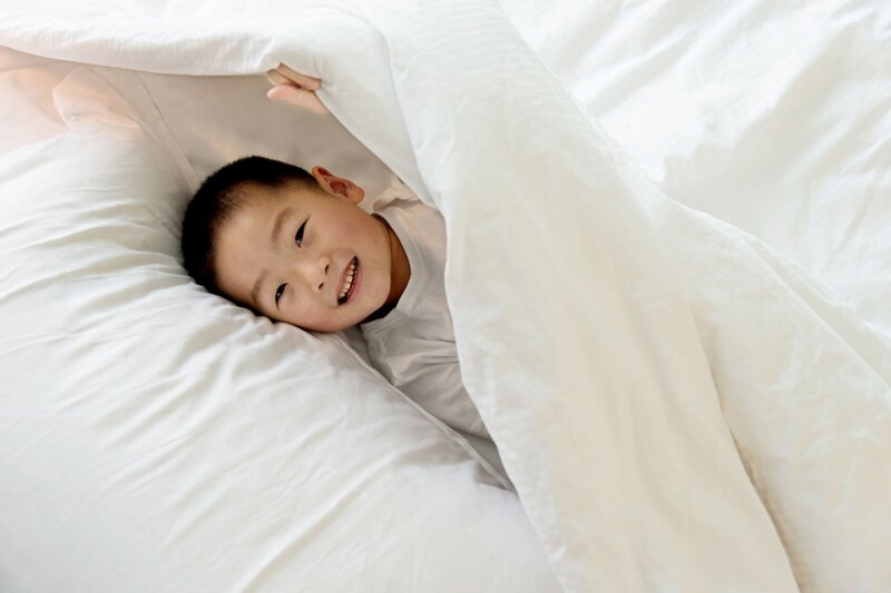 A young asian boy in a bed with white sheets holding the duvet away from his face.