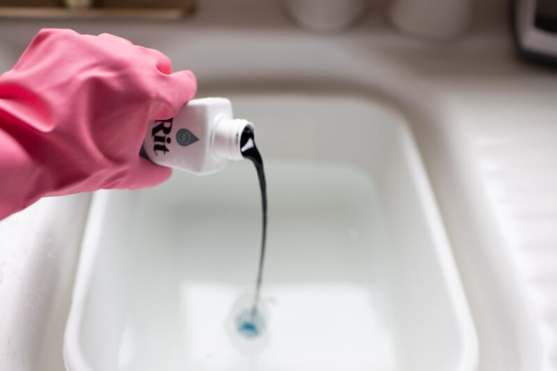 Step two to the ombre tie-dyeing technique for pillowcases. A hand wearing a pink glove pours a bottle of RIT dye into a bucket. 