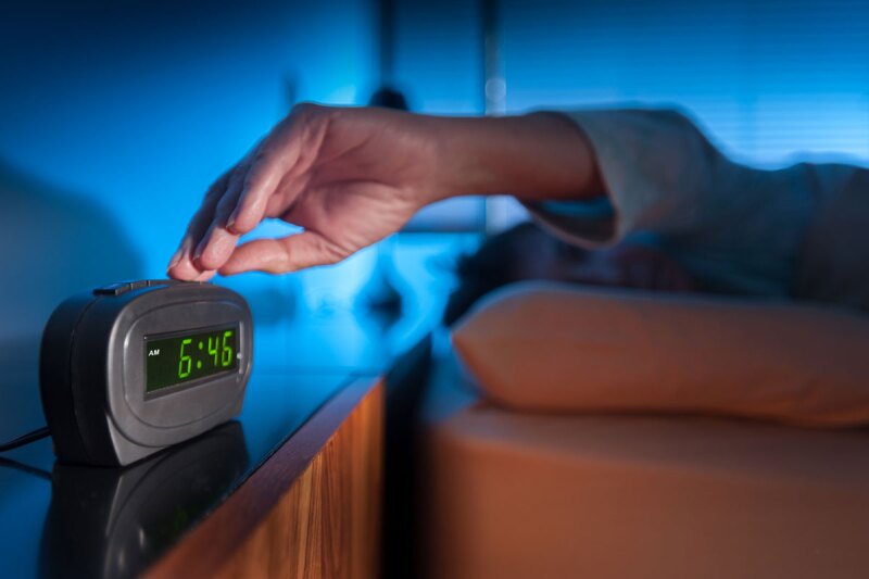 Woman pressing snooze button on early morning digital alarm clock.