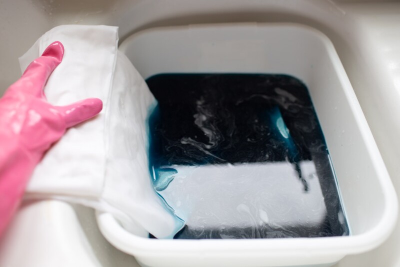 Step three in the DIY ombre tie-dyeing technique. A hand wearing a pink rubber glove submerges a white pillowcase in a bucket of dye. 