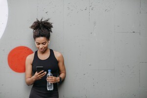 Black woman holding a plastic water bottle and looking at her phone after a workout