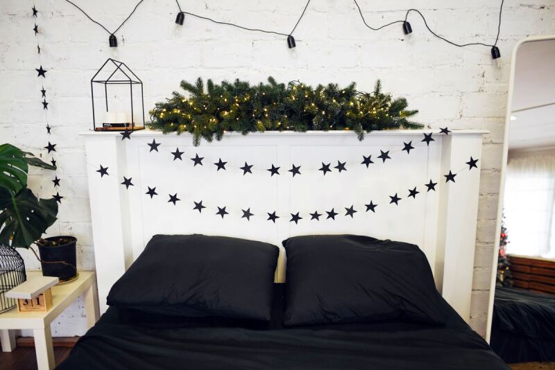 A guest bedroom decorated to celebrate Christmas and the holidays: A bed with two pillows and dark bedding, a string of decorative stars over the headboard, and garland with lights. 