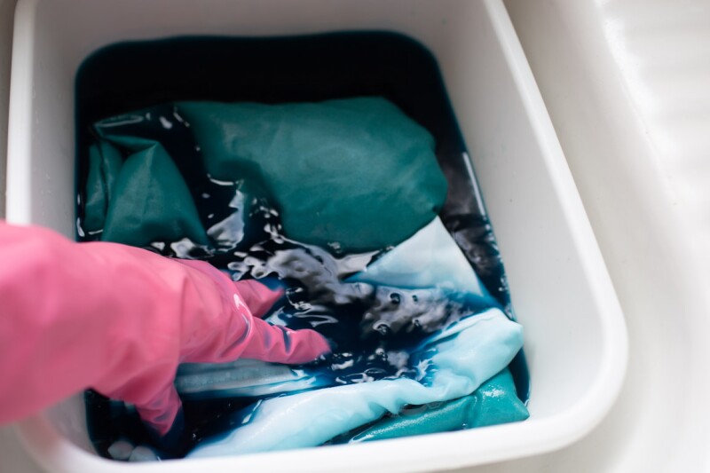 Step four in the DIY ombre tie-dye technique. A hand wearing a pink glove submerges pillowcases in a bucket of water with dye. 