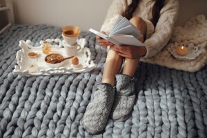 A faceless woman sits on a cozy blanket with a tray of sleep-supporting foods and a candle beside her. She is reading a book. This is part of winter evening routine. 