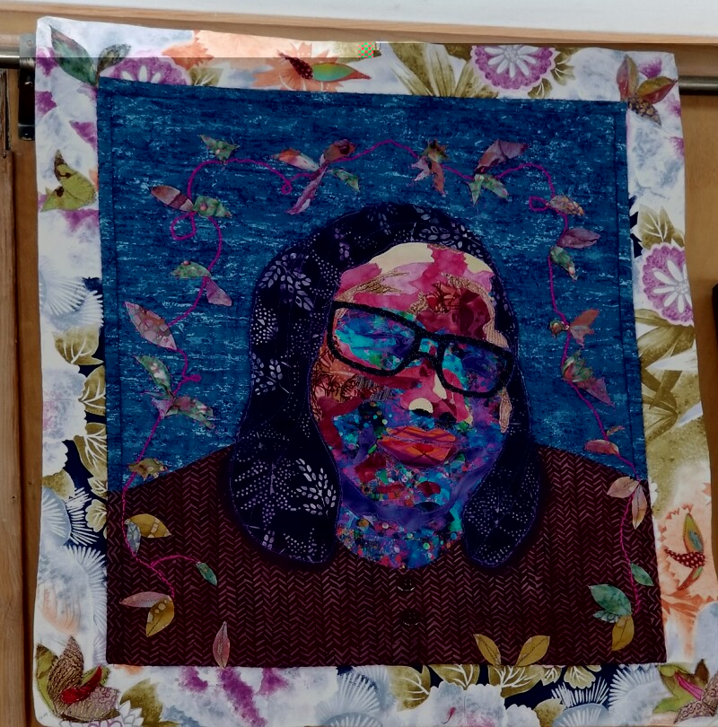 A portrait quilt of a woman wearing glasses, surrounded by abstract designs. This is a quilt by African American quilter Glendora Simonson of the Nubian Heritage Quilters Guild. 