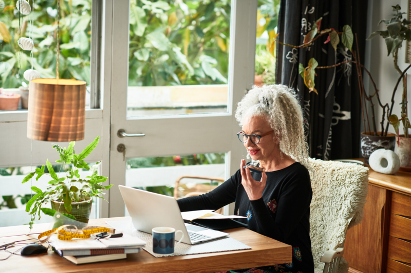 Grey haired female entrepreneur multitasking at home, video conference, speaking and listening on mobile phone, wireless technology.