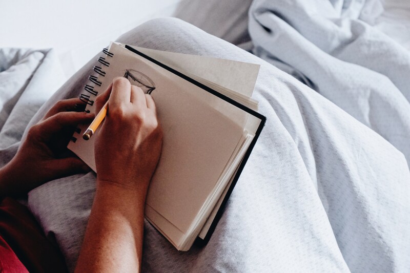 A man sits upright in bed with the covers pulled over his knees. He sketches a doodle into a notebook. This article explores the relationship between sleep deprivation and creativity. 